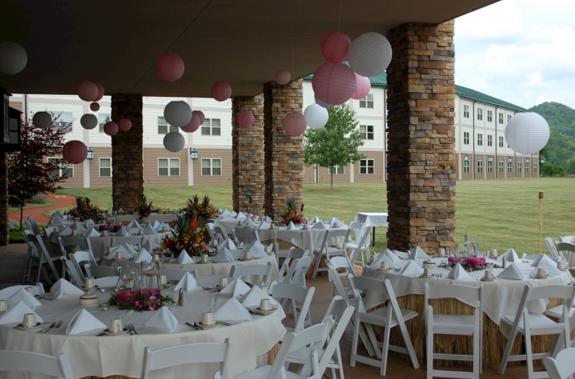 A beautifully decorated outdoor dining area with white chairs, tables, floral centerpieces, and pink and white paper lanterns hanging overhead.