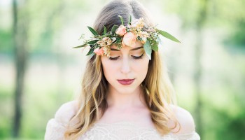 A young woman with blonde hair wears a floral crown with pink roses and greenery, standing outdoors with a blurred green background.