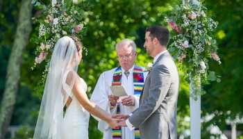 A bride and groom exchange vows under a floral arch with a priest officiating in a sunny outdoor setting. Top it at 140 characters, always ending the sentence.