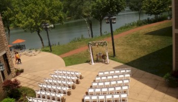 An outdoor wedding setup with rows of white chairs on a stone patio facing a small arch and river beyond, under a sunny blue sky.