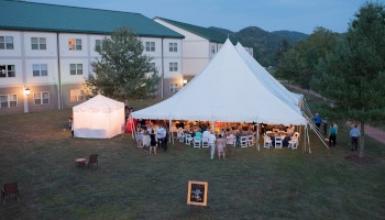 A large white event tent set up on grass outside a building, with guests seated at tables, string lights, and a small chalkboard sign nearby.