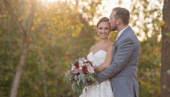 A happy couple in wedding attire outdoors, the groom embracing the bride as she holds a bouquet, with warm sunlight and trees in the background.