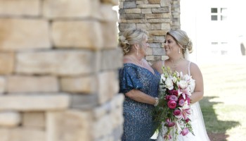 Two brides share a tender moment outdoors, smiling at each other; one in a white wedding gown holds a pink-and-white floral bouquet.