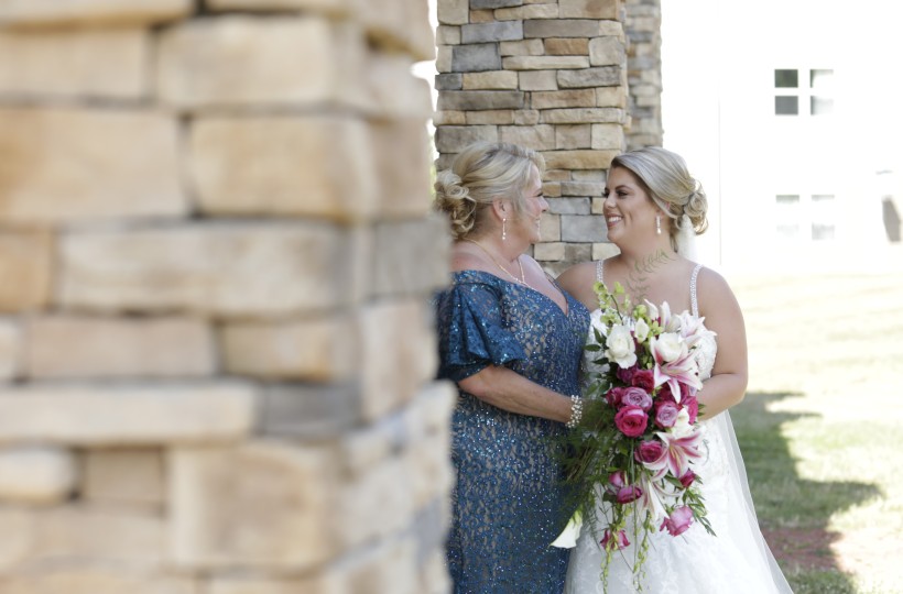 Two brides share a tender moment outdoors, smiling at each other; one in a white wedding gown holds a pink-and-white floral bouquet.