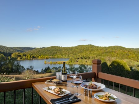 A table set with food and wine overlooks a scenic landscape featuring a lake surrounded by lush greenery, under a clear blue sky.