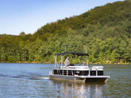 A pontoon boat with a canopy is gliding on a lake with two people aboard, surrounded by lush forest and hills under a clear blue sky.