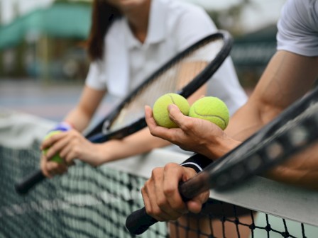 Two people are leaning on a tennis net, holding tennis racquets and tennis balls. The focus is on their hands and tennis equipment.