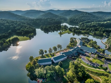 An aerial view of a scenic lakeside resort surrounded by lush green forests and rolling hills under a blue sky with clouds and sunlight reflecting on the water.