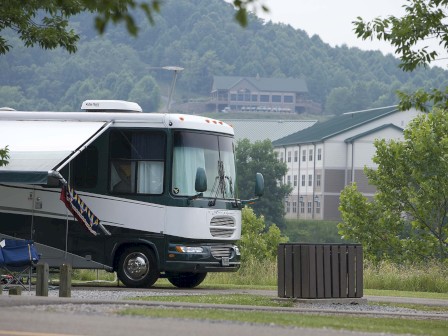 An RV is parked near a scenic area with hills and buildings in the background, and a foldable chair is set up outside.