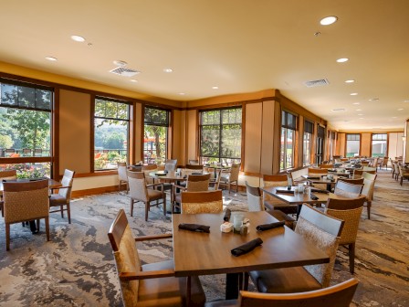 A well-lit restaurant dining area with wooden tables and chairs, set with black napkins, overlooking large windows with an outside view.