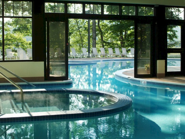 An indoor pool with hot tub and lounge chairs outside, lush green trees, and sunlight shining through large glass windows.