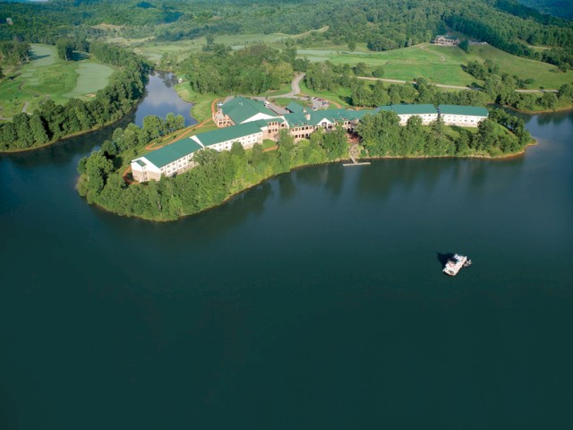 An aerial view shows a large green-roofed building complex on an island surrounded by a lake, with lush forested hills in the background.