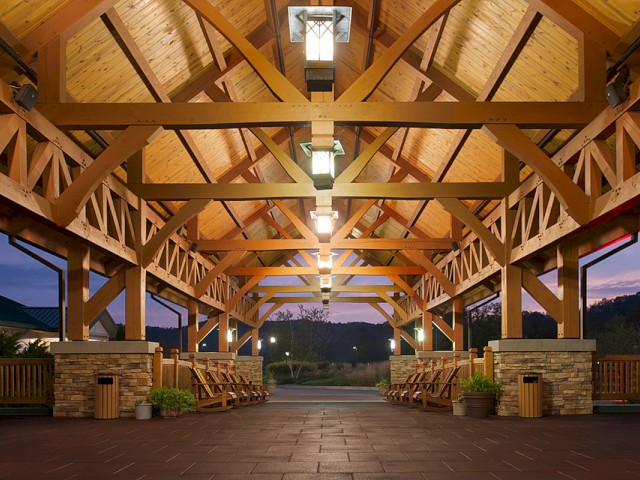 A wooden pavilion with benches, lights, and planters, showcasing warm interior lighting against a dusk sky in a scenic outdoor setting.