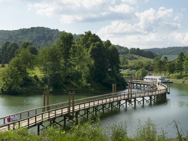 A wooden pier extends over a lake, with people walking and trees, hills, and a building in the background.