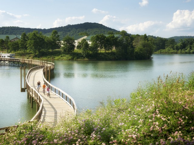 A scenic lakeside view featuring a curved wooden bridge with cyclists, lush greenery, flowers, and distant hills under a partly cloudy sky.