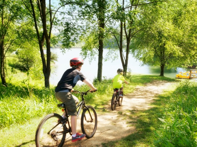 Two children ride bicycles along a sunny, forested path beside a lake, enjoying a peaceful outdoors adventure on a bright day.