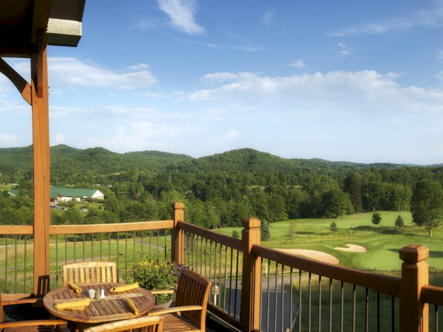 A balcony with a table, chairs, and a view of a lush golf course with green hills under a bright blue sky with some clouds.