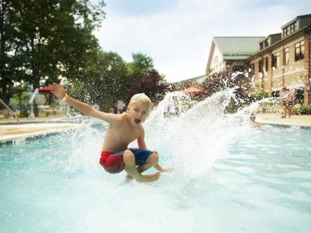 A boy with blonde hair and red swim shorts jumps into a pool, splashing water around on a sunny day at a residential or resort pool area.