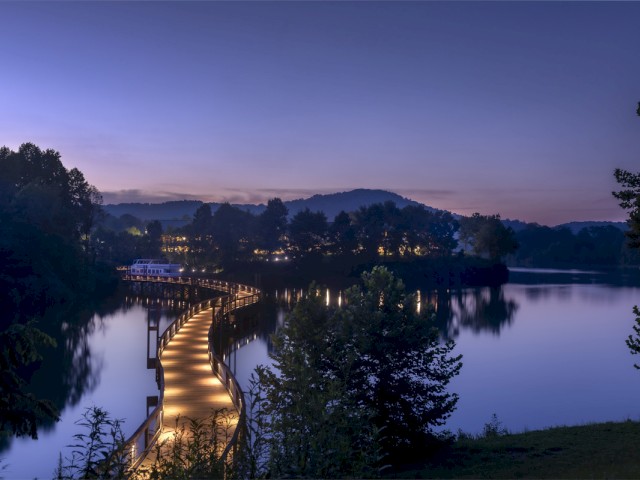 A winding illuminated pathway over a tranquil river at dusk, surrounded by trees and distant hills, creating a peaceful natural scene.