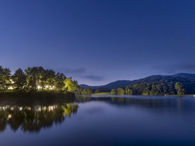 A serene night scene of a river with trees on the shore, mountains in the background, and a clear starry sky reflected in the water.