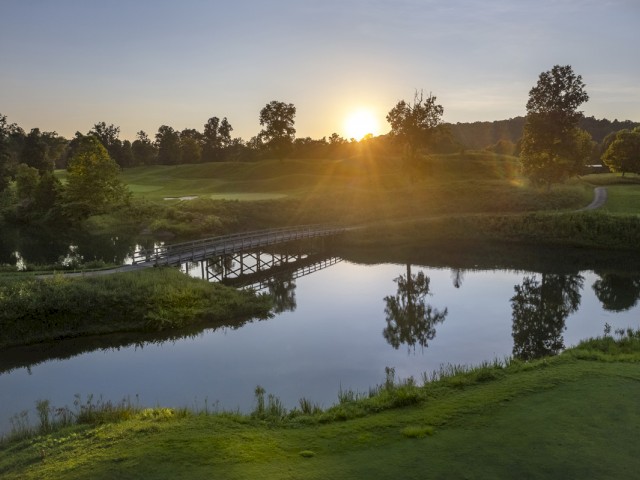 A peaceful golf course at sunset with a bridge over a calm lake, trees, and a winding path in the background.