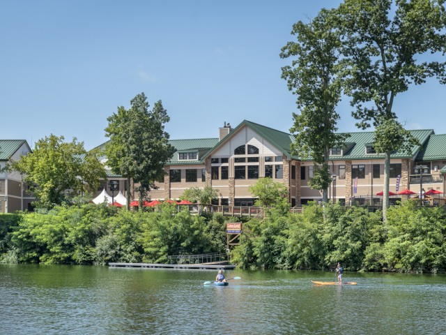 People paddleboarding on a lake with a large house, trees, and a clear blue sky in the background.