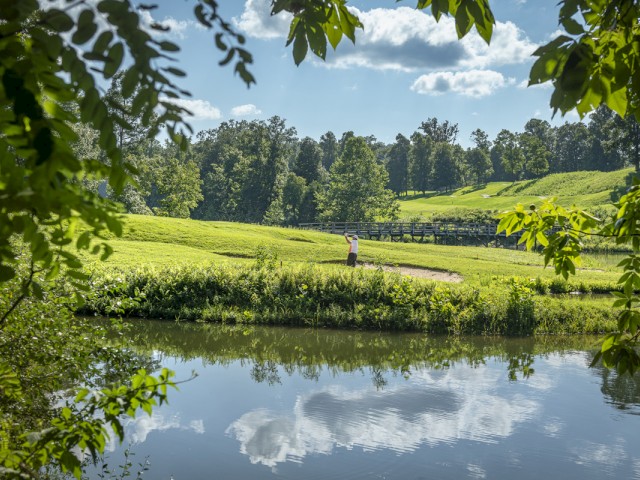 A person stands on a golf course near a body of water, surrounded by lush green trees under a partly cloudy sky.