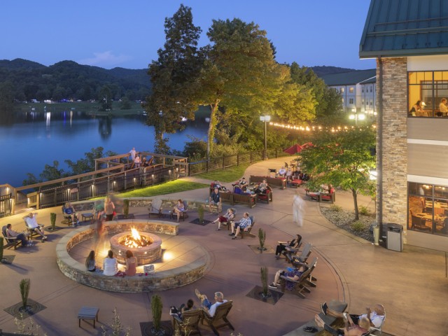 People are relaxing and socializing outside by a fire pit at a waterfront resort during evening, with cozy seating and string lights.