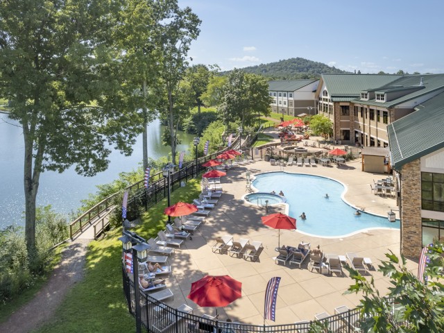 A serene outdoor pool area with red umbrellas, lounge chairs, and people relaxing by a lake with trees and distant hills.
