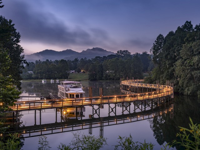 A boat is docked on a calm river with a winding wooden walkway lit by warm lights, surrounded by lush trees and mountains at dusk.