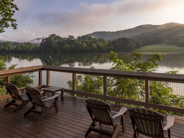 A wooden deck with chairs overlooks a peaceful lake and lush green hills at sunrise or sunset.