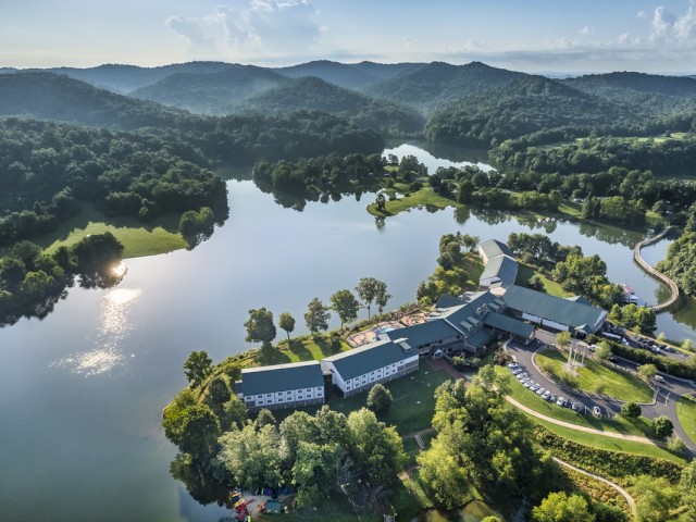A scenic view of a large lakeside retreat surrounded by lush green mountains, with buildings, parking, and a winding driveway visible.