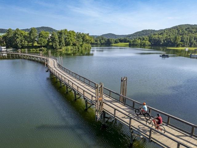Two children ride bikes on a wooden bridge over a calm lake with green trees and hills in the background on a sunny day.