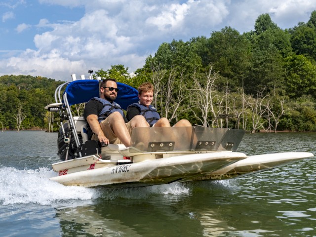 Two men sit on a boat with a canopy, riding on a lake surrounded by green trees and a partly cloudy sky.