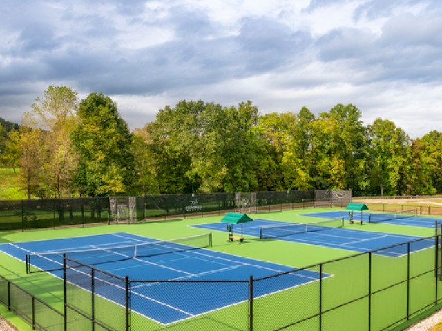 A bright outdoor tennis court with blue surface, surrounded by a fence, two umbrellas, and lush green trees under cloudy skies.