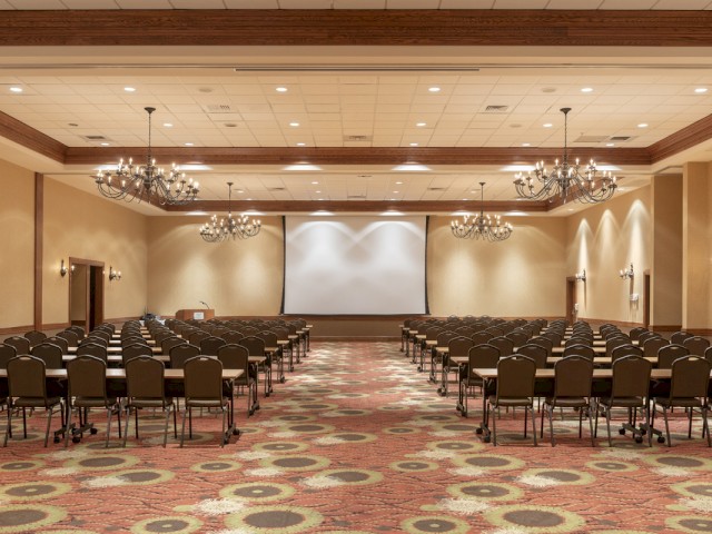 This is an empty conference room with rows of tables, chairs, chandeliers, and a large screen at the front, ready for a presentation.