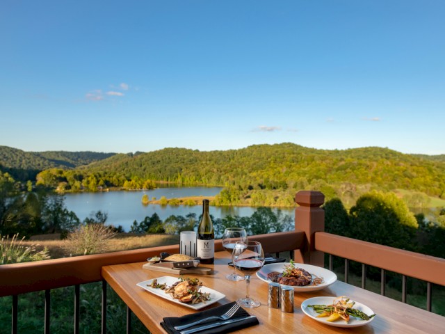 A dinner setup with wine, steak, vegetables, and desserts on a wooden table overlooking a lake and green hills during sunset.