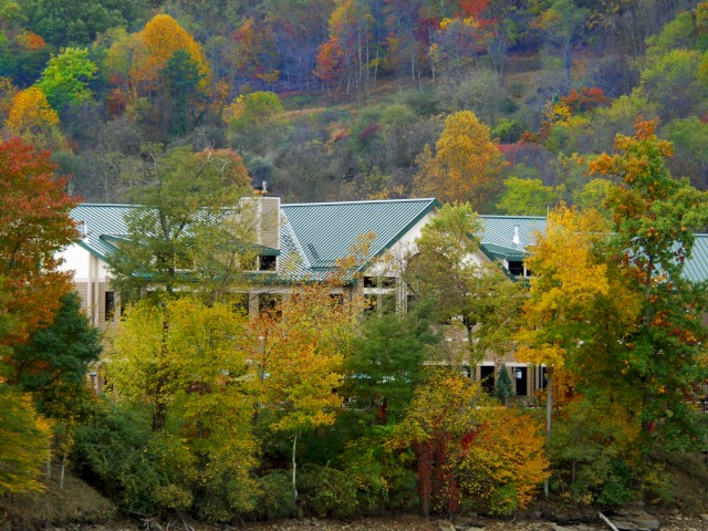 A house with a green roof is surrounded by colorful autumn trees. The hillside in the background displays fall foliage.