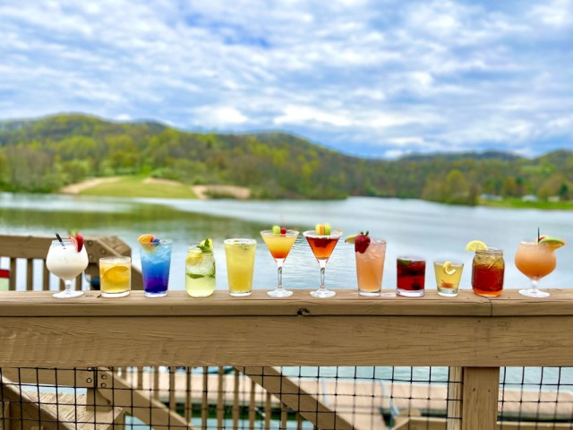 A variety of colorful cocktails are lined up on a wooden railing by a lake with hills and a cloudy sky in the background.