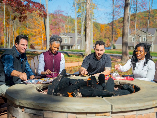A diverse group of five people are roasting marshmallows around a backyard fire pit on a sunny autumn day.