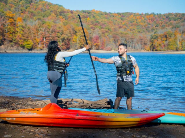 Two people near the water's edge are engaging in a playful exchange with paddles, with a kayak and a paddleboard on the shore in autumn.