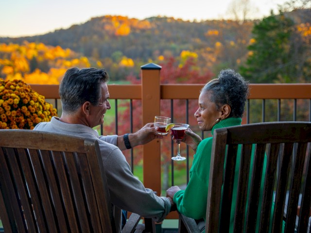 A man and woman share drinks on a balcony with fall foliage in the background amid a warm sunset.