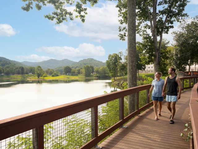 A couple is walking hand in hand on a wooden trail beside a calm lake surrounded by lush trees and distant green mountains under a partly cloudy sky.