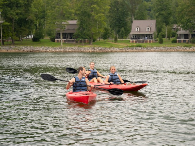 Three people are kayaking on a lake near a residential area with trees and houses in the background. They are smiling and enjoying the activity.