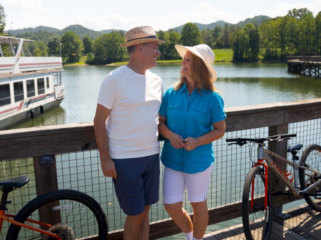 A couple is smiling and talking on a sunny dock by the water, with bikes, a boat, and trees in the background.