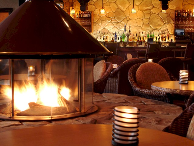 A cozy bar with a lit fireplace, round tables, cushioned chairs, and a well-stocked bar in a warm, inviting ambiance.