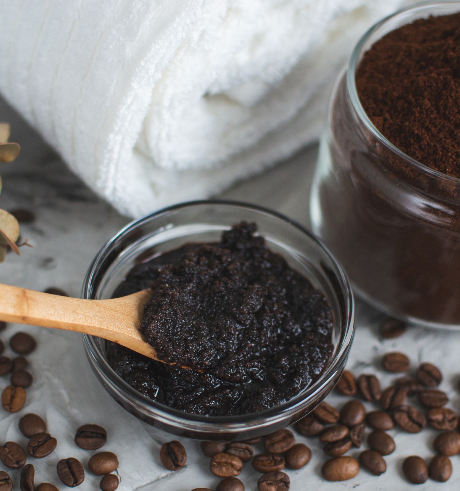 The image shows a jar of coffee grounds, a bowl of scrub with a wooden spoon, coffee beans, eucalyptus leaves, and a white towel.