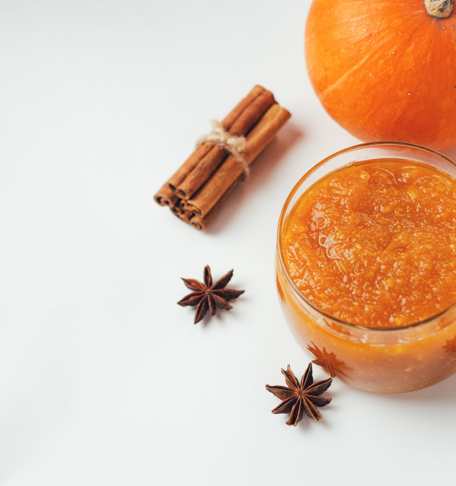 The image shows a jar of pumpkin jam, a small pumpkin, a bundle of cinnamon sticks, and star anise on a white background.