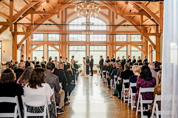 A wedding ceremony in a wooden hall with a chandelier, guests seated in rows, couple at the altar, sunlit arched windows behind them.