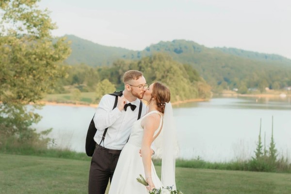 A bride in a white gown and a groom in a black tux share a kiss by a lakeside, holding a bouquet, with a scenic grassy backdrop.